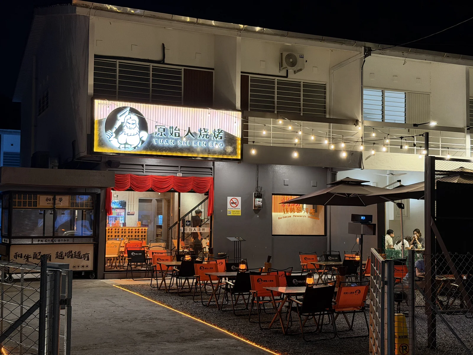 The Tanjung Bungah shopfront at night with its lit caveman signboard and garden tables